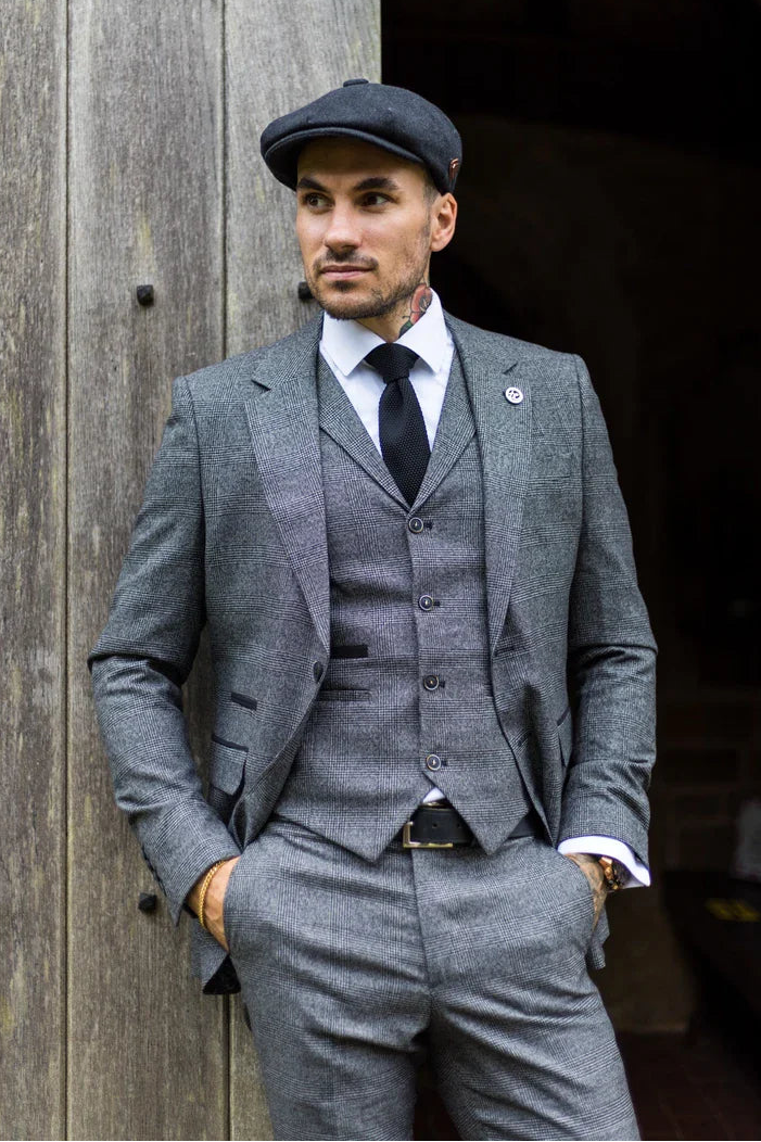 Man in a gray suit and cap standing against a wooden wall.